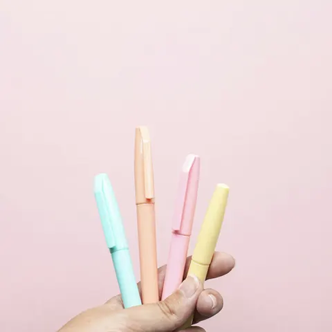 A hand holding four pastel highlighter pens against a pink wall