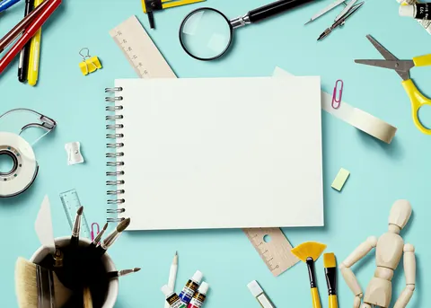 A blank spiral sketch pad surrounded by art supplies on a blue desk