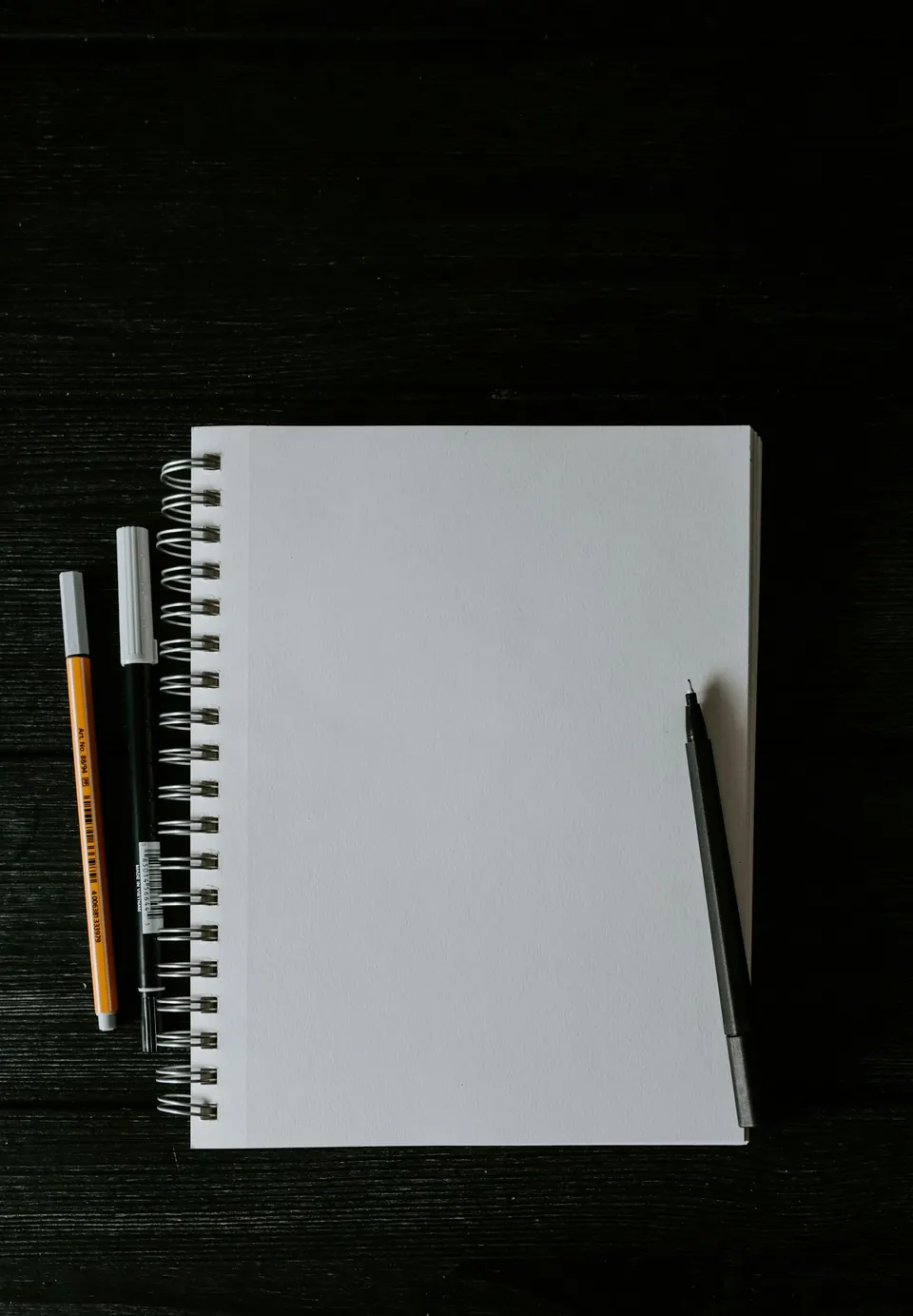 A spiral notebook with a pencil and a pen on a dark desk